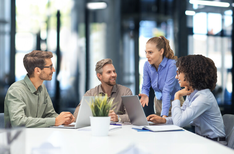 Vier glücklich aussehende Menschen in einer Geschäftsbesprechung. Eine Frau steht, eine andere Frau und zwei Männer sitzen dafür im Kreis an einem Tisch mit Laptops vor sich.Foto: (c) ty / AdobeStock